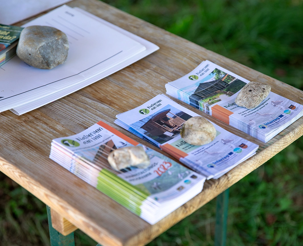 Informational flyers and brochures on a wooden table outdoors, weighted down with stones.