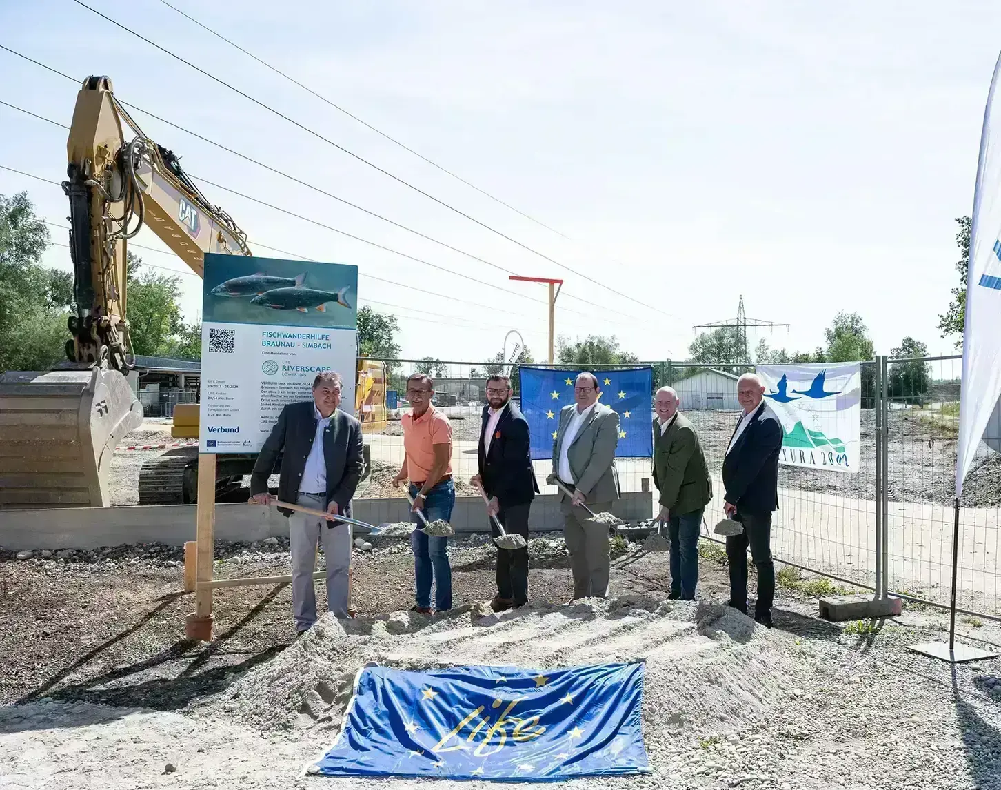 Group of individuals at a groundbreaking ceremony at a construction site with an excavator and informational signs in the background.