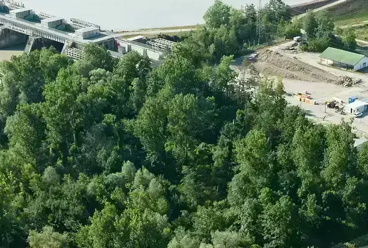 Luftaufnahme eines Wasserkraftwerks am Fluss mit einem Waldgebiet im Vordergrund und landwirtschaftlichen Feldern im Hintergrund.