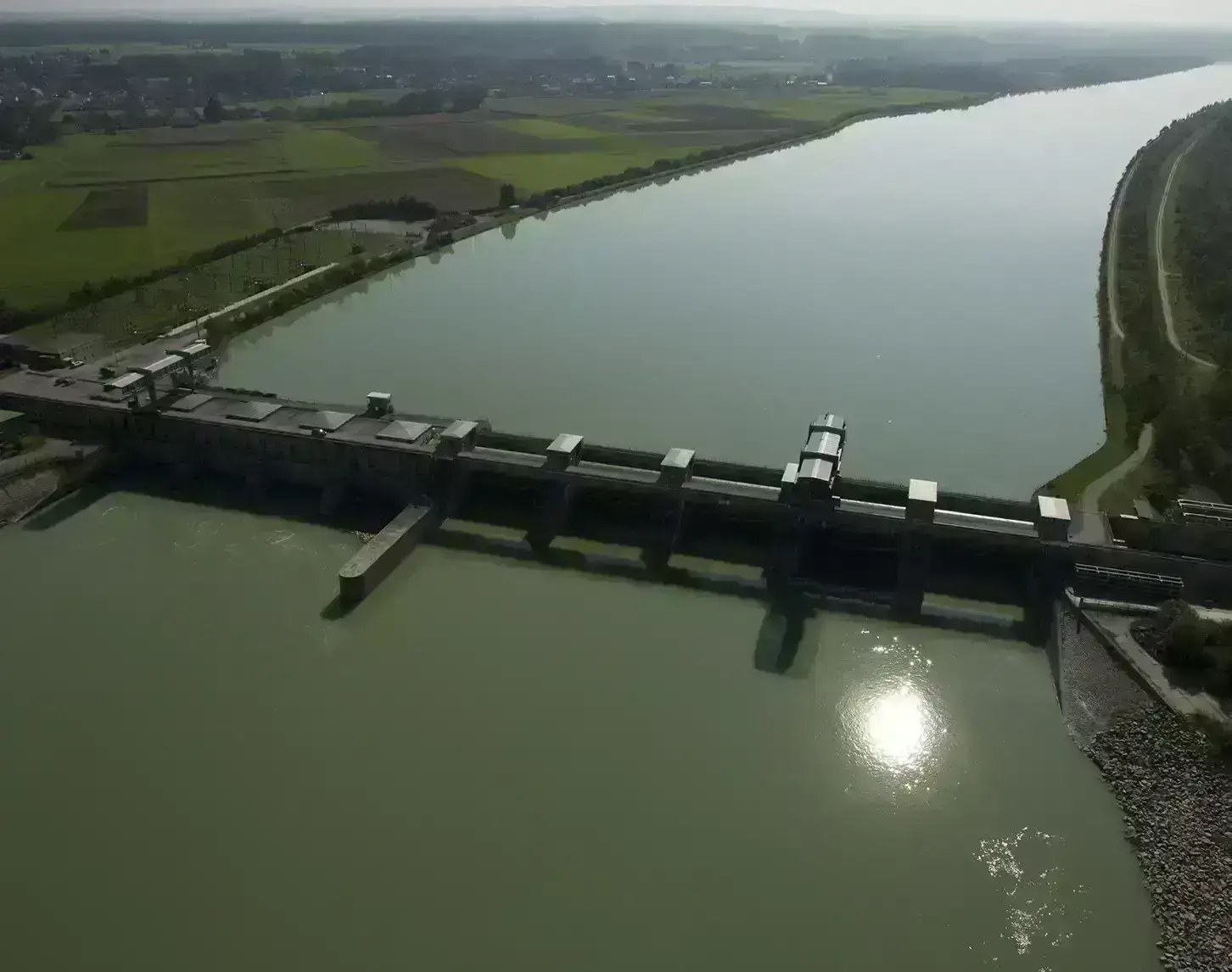Luftaufnahme eines Laufkraftwerks an einem breiten Fluss, umgeben von landwirtschaftliche Feldern an einem sonnigen Tag.