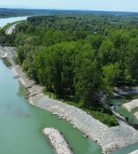 Luftaufnahme eines Kraftwerks an einem Fluss mit aufgestautem Wasser, angrenzendem Waldgebiet und Wanderwegen bei klarem Wetter.