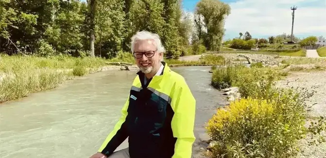 Individual in a yellow jacket by the bank of a flowing river in a green landscape with trees and shrubs in the background.